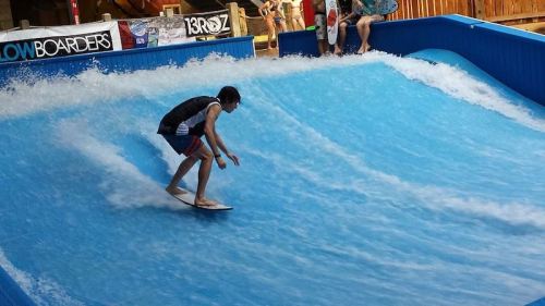 A person surfing on a blue wave in a flow rider attraction, with spectators in the background.