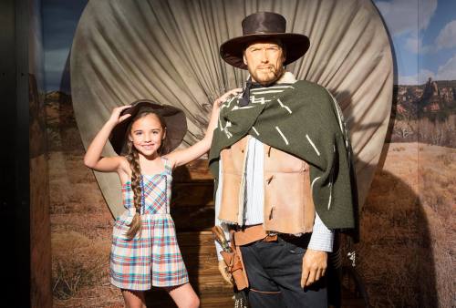 A girl in a plaid dress poses with a cowboy figure in front of a western-themed backdrop.