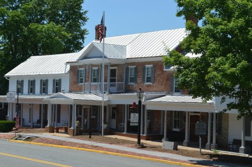 Historic buildings with white metal roofs and porches, featuring American flags, along a tree-lined street.