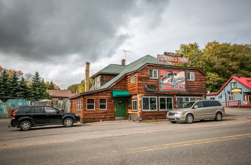 A rustic wooden building with a green roof, featuring a sign and parked cars in front, under a cloudy sky.