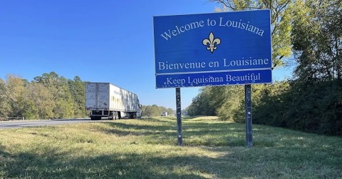 A welcome sign for Louisiana, featuring a fleur-de-lis, with a truck passing by on a clear day.