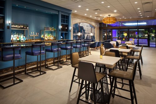Modern bar area with high tables and stools, blue walls, and a well-stocked bar in a stylish restaurant setting.