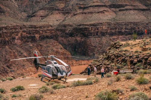 A helicopter parked on rocky terrain near a canyon, with people exploring the area in the background.
