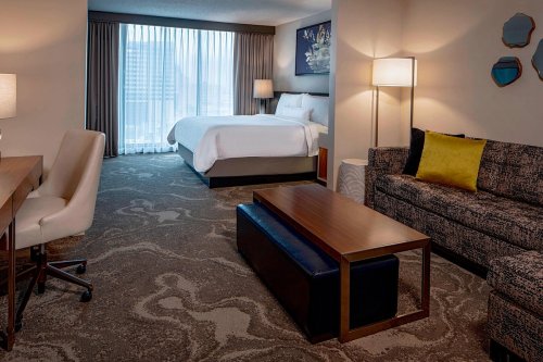 A modern hotel room featuring a bed, sofa, desk, and large window with city views.