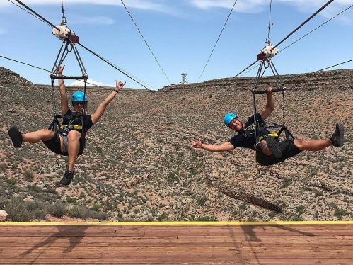 Two people ziplining over a rocky landscape, smiling and posing with their hands in the air.