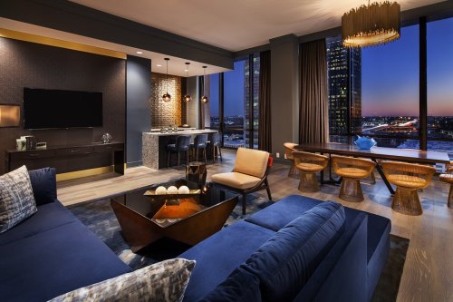 Modern living room with a blue sofa, wooden dining table, and large windows showcasing a city skyline at sunset.