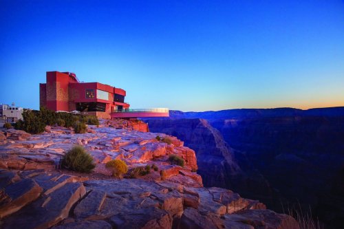A modern building perched on a rocky ledge overlooking the Grand Canyon at sunset, with vibrant blue and orange hues.