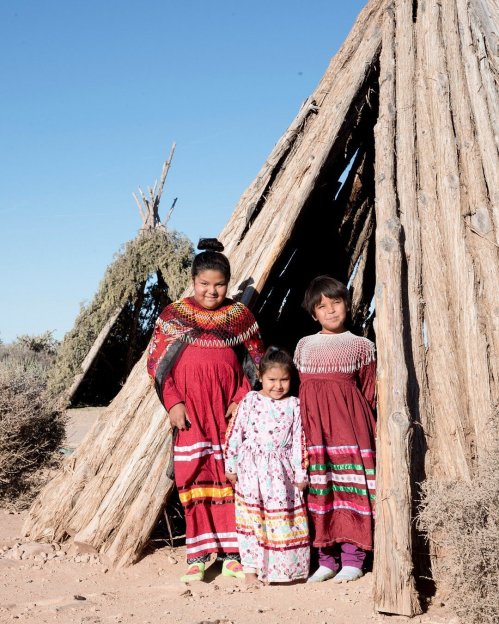 Three young girls in traditional dresses stand outside a wooden structure in a desert landscape.