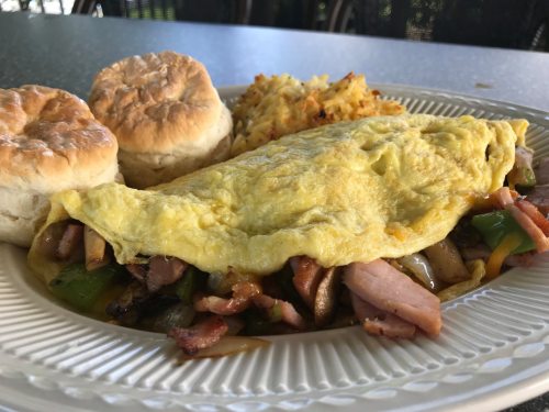 A plate featuring a fluffy omelet filled with vegetables and meats, served with biscuits and hash browns.