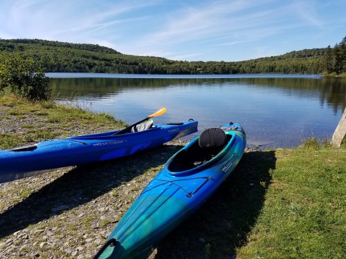 Two kayaks sit on the shore of a calm lake, surrounded by green hills and a clear blue sky.