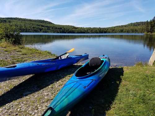 Two kayaks sit on the shore of a calm lake, surrounded by green hills and a clear blue sky.