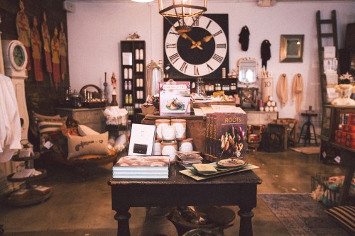 A cozy shop interior featuring a large clock, shelves of products, and a table with books and decorative items.