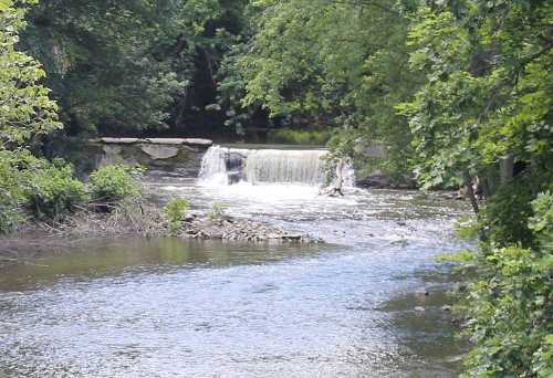 A serene river scene featuring a small waterfall surrounded by lush green trees.