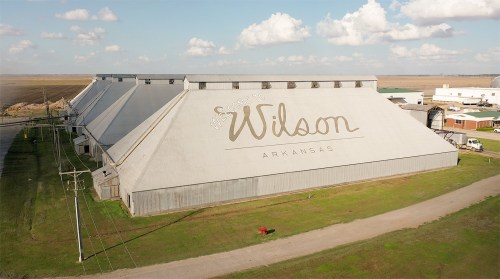 Aerial view of large warehouses with "Welcome to Wilson, Arkansas" painted on the side, surrounded by open fields.