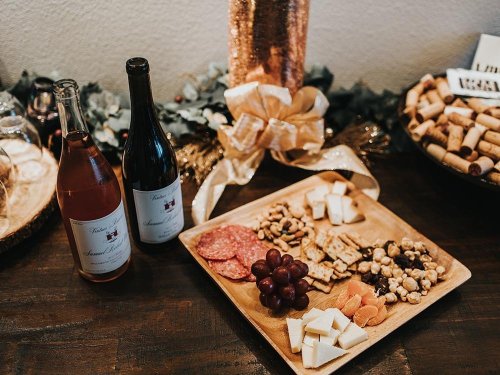 A wooden platter with assorted cheeses, meats, nuts, and grapes, alongside two bottles of wine and decorative elements.