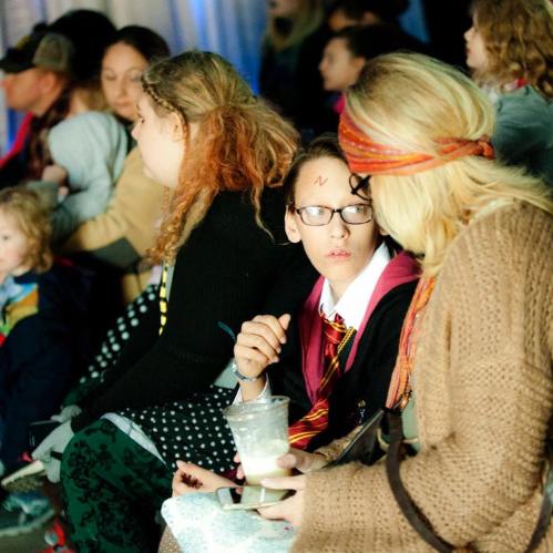 A child in glasses and a Harry Potter costume sits with a woman, both engaged in conversation at a crowded event.