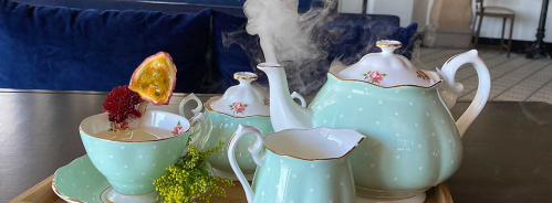 A pastel blue tea set with floral accents, steam rising from a teapot, and a decorative cup on a wooden tray.
