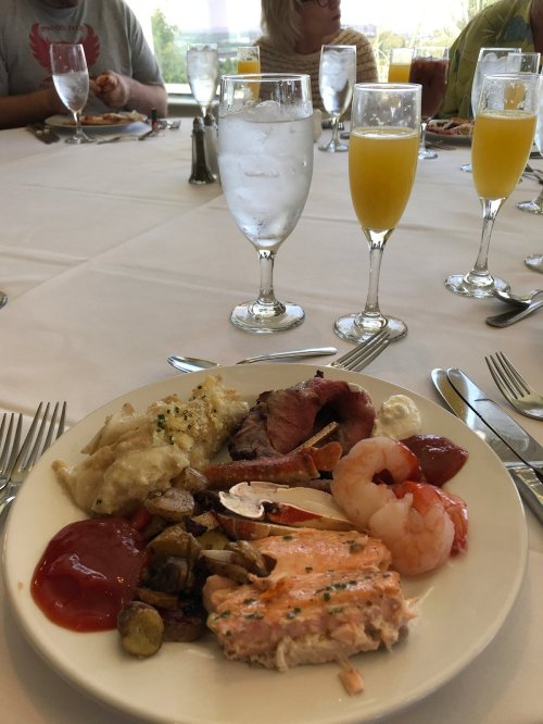 A plate of food featuring shrimp, salmon, roast beef, potatoes, and a side of sauce, with drinks and diners in the background.