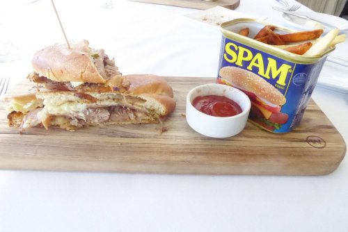 A wooden board with a sandwich stacked with meat and cheese, alongside a can of Spam and a small bowl of ketchup.