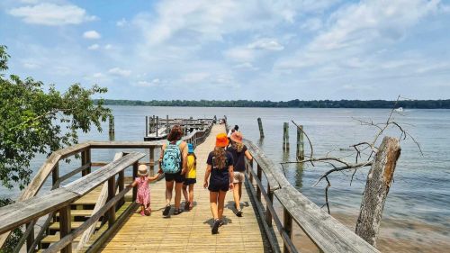 A group of people walking along a wooden pier by a calm river under a partly cloudy sky.
