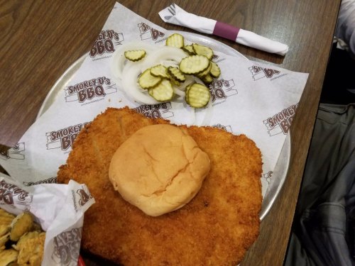 A plate with a large breaded chicken sandwich, pickles, and onions on paper with a BBQ logo, alongside a fork and knife.