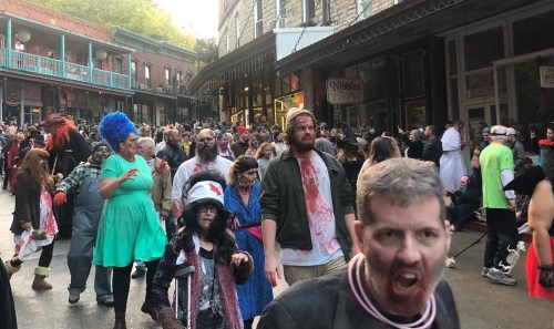 A crowd of people in zombie costumes walking down a street during a Halloween event, with colorful outfits and makeup.