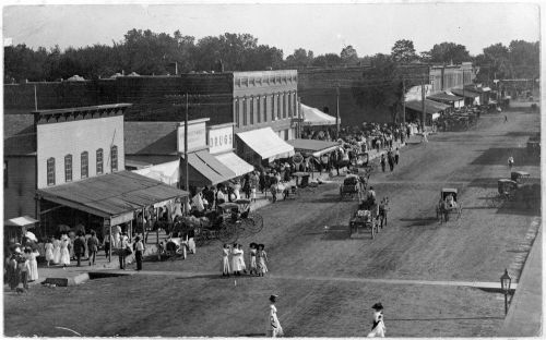 A historic street scene with horse-drawn carriages, people walking, and storefronts under awnings on a sunny day.