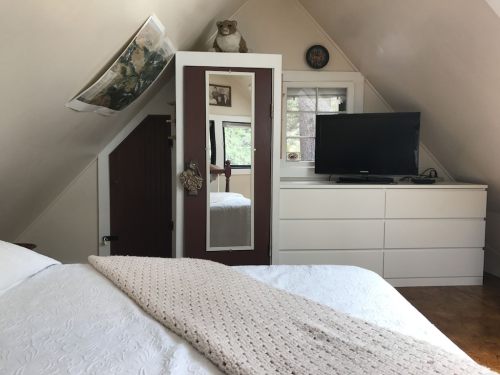 Cozy attic bedroom with a bed, dresser, TV, and a mirror on the wall, featuring warm lighting and a decorative touch.