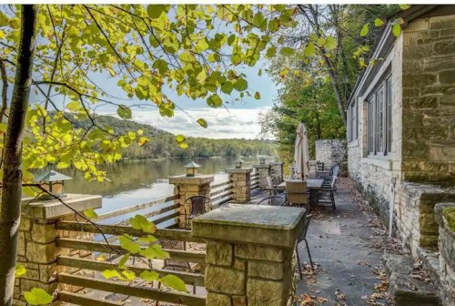 A serene riverside patio with stone walls, surrounded by green trees and a clear blue sky.