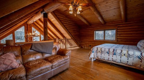 Cozy wooden attic bedroom with a bed, leather sofa, and large windows, featuring rustic decor and warm lighting.