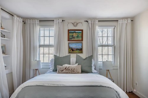Cozy bedroom with a bed, decorative pillows, two lamps, and large windows with white curtains.