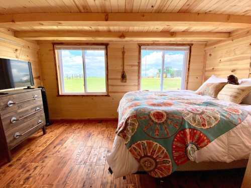 Cozy bedroom with wooden walls, a large bed, colorful quilt, and windows overlooking a green landscape.