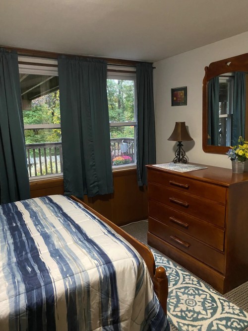 Cozy bedroom with a wooden dresser, a lamp, and large windows with green curtains overlooking a garden.
