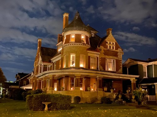 A grand, historic house with a turret, illuminated windows, and a cloudy night sky in the background.