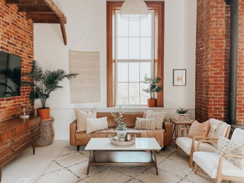 Cozy living room with brick walls, plants, a brown sofa, and a light rug, featuring natural decor and a large window.