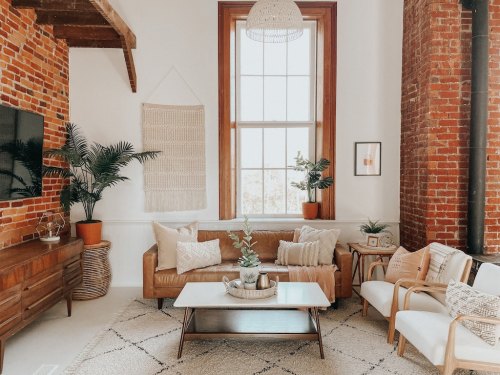 Cozy living room with a brown sofa, white chairs, plants, and a large window, featuring brick walls and a stylish rug.