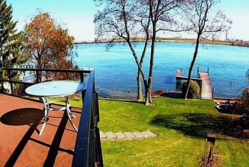 A scenic view of a lake from a balcony, featuring a table, green lawn, trees, and a wooden dock.