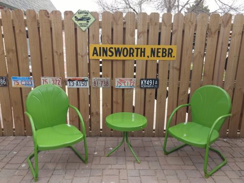 Two green chairs and a small table in front of a wooden fence with a sign reading "AINSWORTH, NEBR." and old license plates.