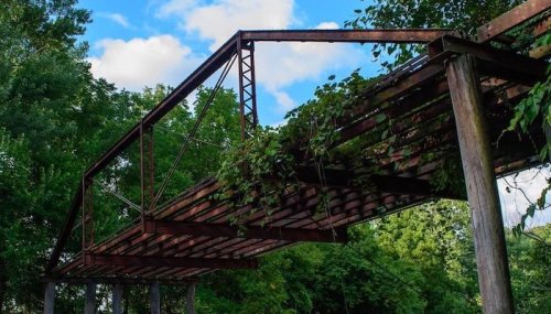 An old, rusted bridge covered in greenery, surrounded by trees and a blue sky with clouds.