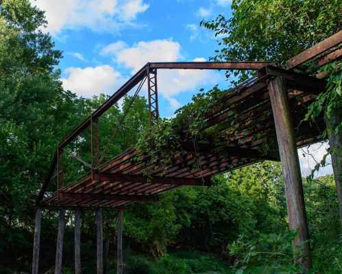 An old, rusted bridge partially covered in greenery, set against a blue sky with scattered clouds.