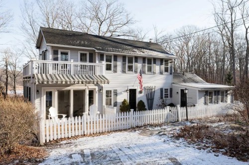 A two-story white house with a porch, surrounded by trees and a white picket fence, in a snowy landscape.