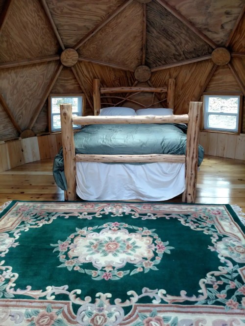 Cozy interior of a geodesic dome with a wooden bed, green bedding, and a decorative rug on the wooden floor.