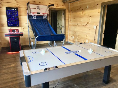 A game room featuring an air hockey table, basketball arcade, and a retro arcade machine against a wooden wall.