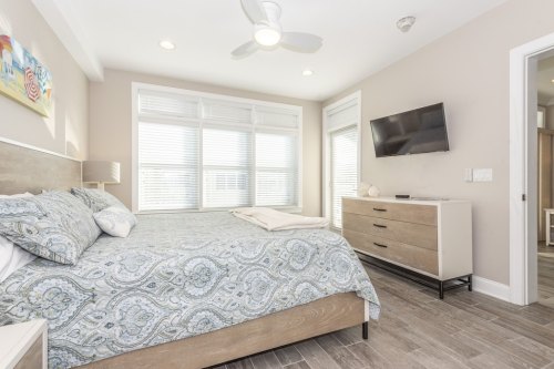 Bright bedroom featuring a queen bed, dresser, TV, and large windows with natural light. Modern decor and neutral tones.