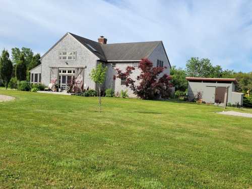 A charming gray house with a sloped roof, surrounded by green grass and trees, with a small shed nearby.