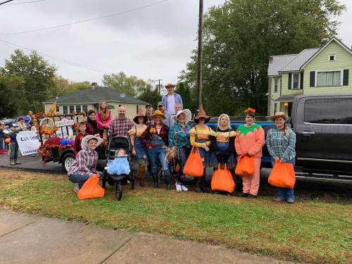 A group of people in costumes poses for a photo, holding orange bags, with a decorated truck in the background.