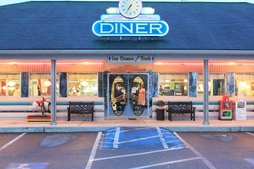 A retro diner with neon signage, featuring surfboards and outdoor seating, set in a parking lot at dusk.