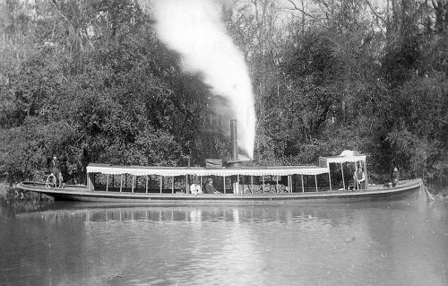 A historic steamboat on a river, with smoke billowing from its stack and surrounded by lush greenery.