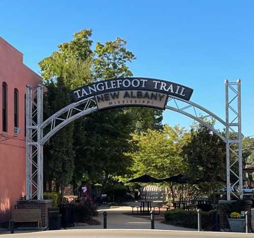 Archway sign for Tanglefoot Trail in New Albany, Mississippi, surrounded by trees and outdoor seating. Clear blue sky.