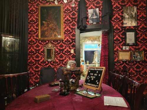 A dimly lit room with red damask wallpaper, a round table, and various framed artworks and decor items.